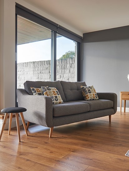 Mid century modern sofa in grey fabric with bright scatter cushions shown against a dark wall, wooden floor and a big window